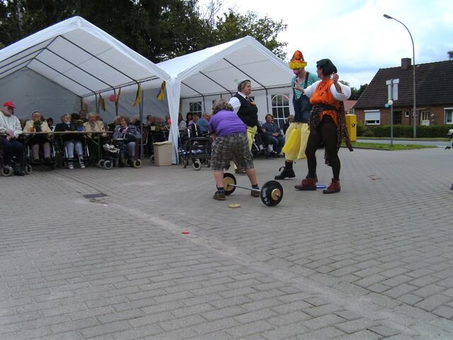 Eine humorvolle Veranstaltung im Freien, bei der zwei verkleidete Clowns mit bunten Kostümen auf einer kleinen Bühne auftreten. Im Hintergrund sitzen ältere Personen in Rollstühlen und beobachten begeistert die Darbietung.