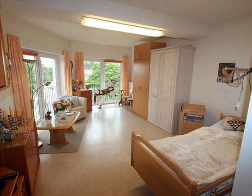 Ein heller, geräumiger Raum mit einem Bett, einem Tisch und Stühlen. Auf dem Tisch steht eine Blumenvase. Große Fenster bieten einen Blick nach draußen. Ein Kleiderschrank und weitere Möbel ergänzen den Raum. Die Atmosphäre wirkt freundlich und einladend.