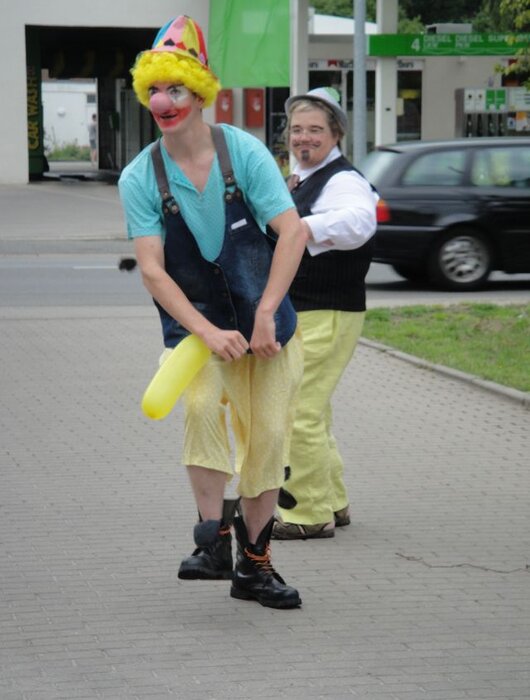 Zwei Clowns stehen auf einem Gehweg. Der vordere Clown trägt eine bunte Perücke, ein blaues Oberteil und gelbe Hosen. Er hält einen langen gelben Luftballon. Der zweite Clown folgt ihm, gekleidet in eine schwarze Weste und weiße Hemden, mit einem fröhlichen Ausdruck.