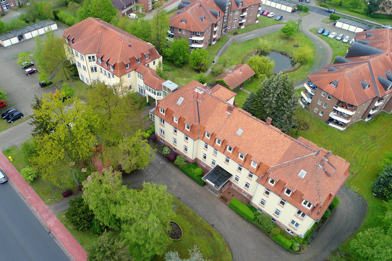 Luftaufnahme eines Gebäudekomplexes mit mehreren Wohnhäusern und gepflegten Grünanlagen. Auf dem Gelände sind Bäume, Sträucher und ein kleiner Teich sichtbar. Die Dächer sind rot und der Bereich ist umgeben von Straßen und Parkplätzen.