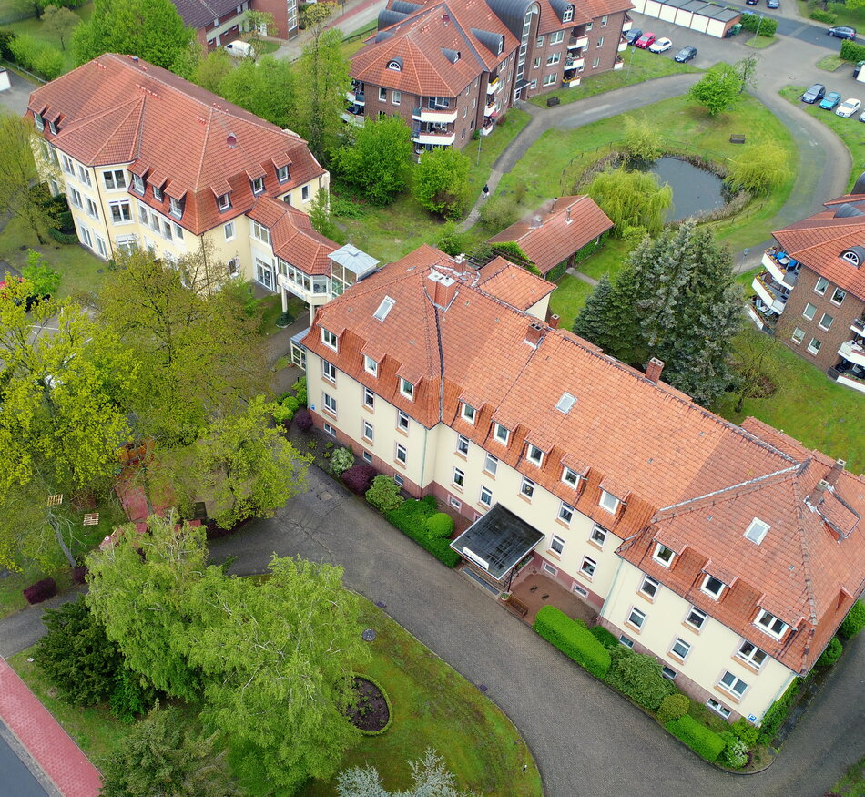 Luftaufnahme eines Gebäudekomplexes mit mehreren Wohnhäusern und gepflegten Grünanlagen. Auf dem Gelände sind Bäume, Sträucher und ein kleiner Teich sichtbar. Die Dächer sind rot und der Bereich ist umgeben von Straßen und Parkplätzen.
