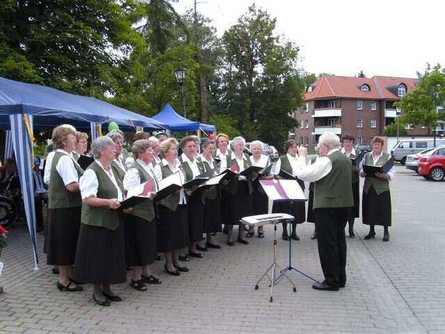 Eine Gruppe von etwa 20 Sängerinnen in traditionellen Trachten steht im Freien und singt. Ein Dirigent leitet den Chor. Im Hintergrund sind Bäume und einige Gebäude zu sehen. Es ist ein sonniger Tag.