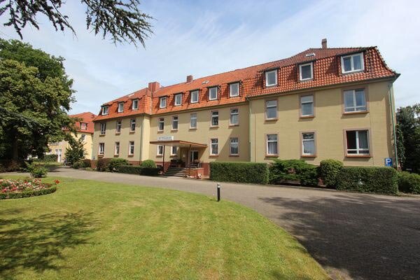 Ein mehrstöckiges Gebäude mit gelber Fassade und roten Dachziegeln. Es verfügt über mehrere Fenster und einen Eingang mit Treppe. Vor dem Gebäude erstreckt sich eine grüne Rasenfläche, umgeben von Bäumen und einem kleinen Parkplatz.