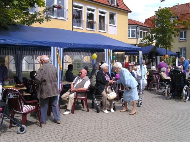 Eine Gruppe älterer Menschen sitzt und steht unter blauen Zelten im Freien. Einige haben Rollatoren oder Rollstühle. Die Atmosphäre wirkt gesellig, mit mehreren Personen, die miteinander sprechen und lachen, während sie die Tageszeit im Freien genießen.