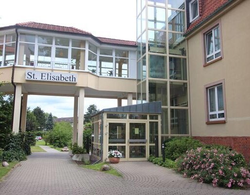 Die Aufnahme zeigt den Eingangsbereich des Gebäudes "St. Elisabeth". Es ist ein mehrstöckiges Haus mit großen Fenstern und einem Aufzug. Der Eingangsbereich ist von Pflanzen und Wegen umgeben, die zum Parkplatz führen.